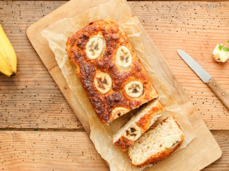 Angeschnittener Bananenkuchen auf Backpapier von oben fotografiert. Daneben liegen ein Messer und Bananen.