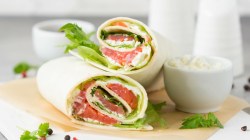 2 Wraps mit Lachs auf einem Brett, daneben frische Petersilie, Pfefferkörner und eine kleine Schüssel mit Frischkäse.