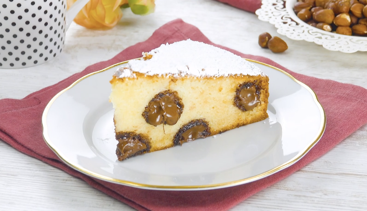 Ein Stück Kuchen mit Nutella-Füllung auf einem weißen Teller. Haselnüsse und eine Tasse im Hintergrund. 