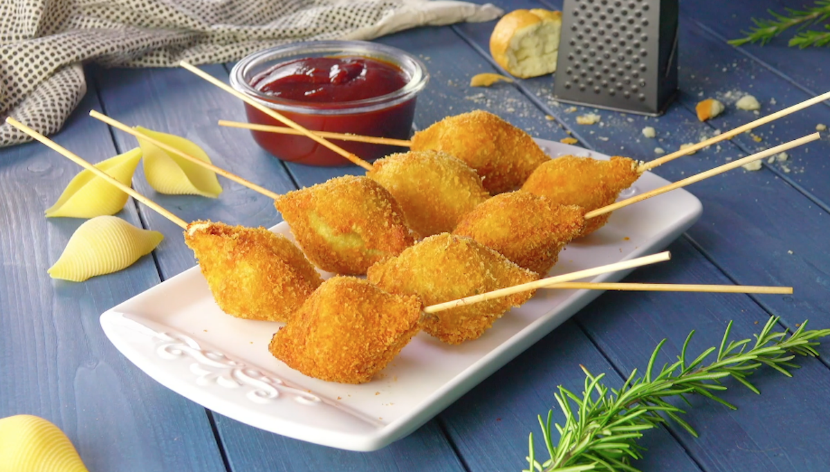 8 herzhafte Lollis auf einer länglichen Servierplatte mit einer Küchenreibe, einem Schälchen mit Ketchup und weiteren Zutaten im Hintergrund.