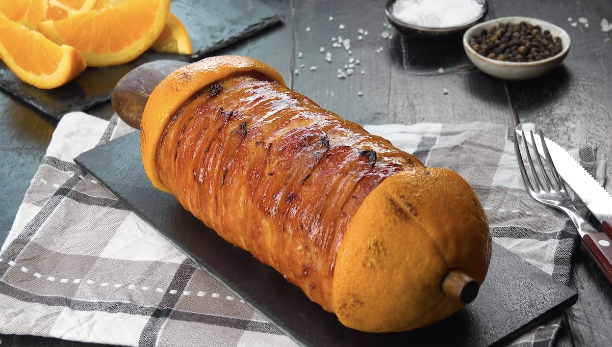 Ein Cordon bleu am Spieß vom Grill neben Besteck, Schälchen mit Salz und Pfeffer und Orangenspalten.