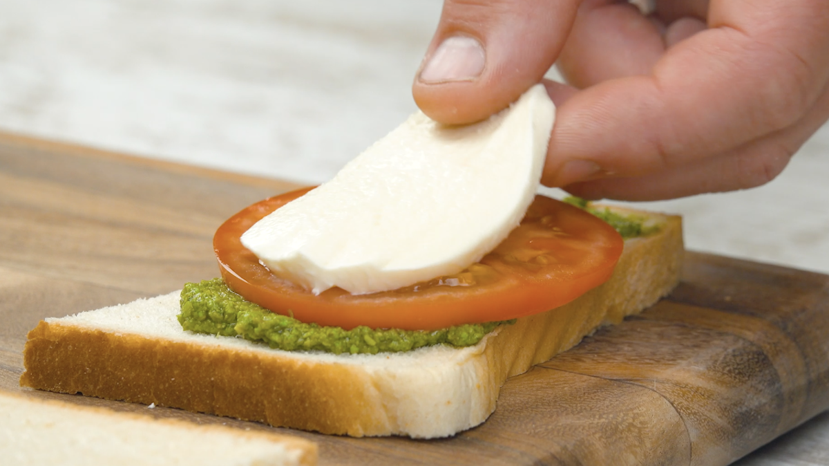 Die grÃ¶ÃŸere ToastbrothÃ¤lfte wird mit Pesto und einer Tomatenscheibe und einer Scheibe Mozzarella belegt