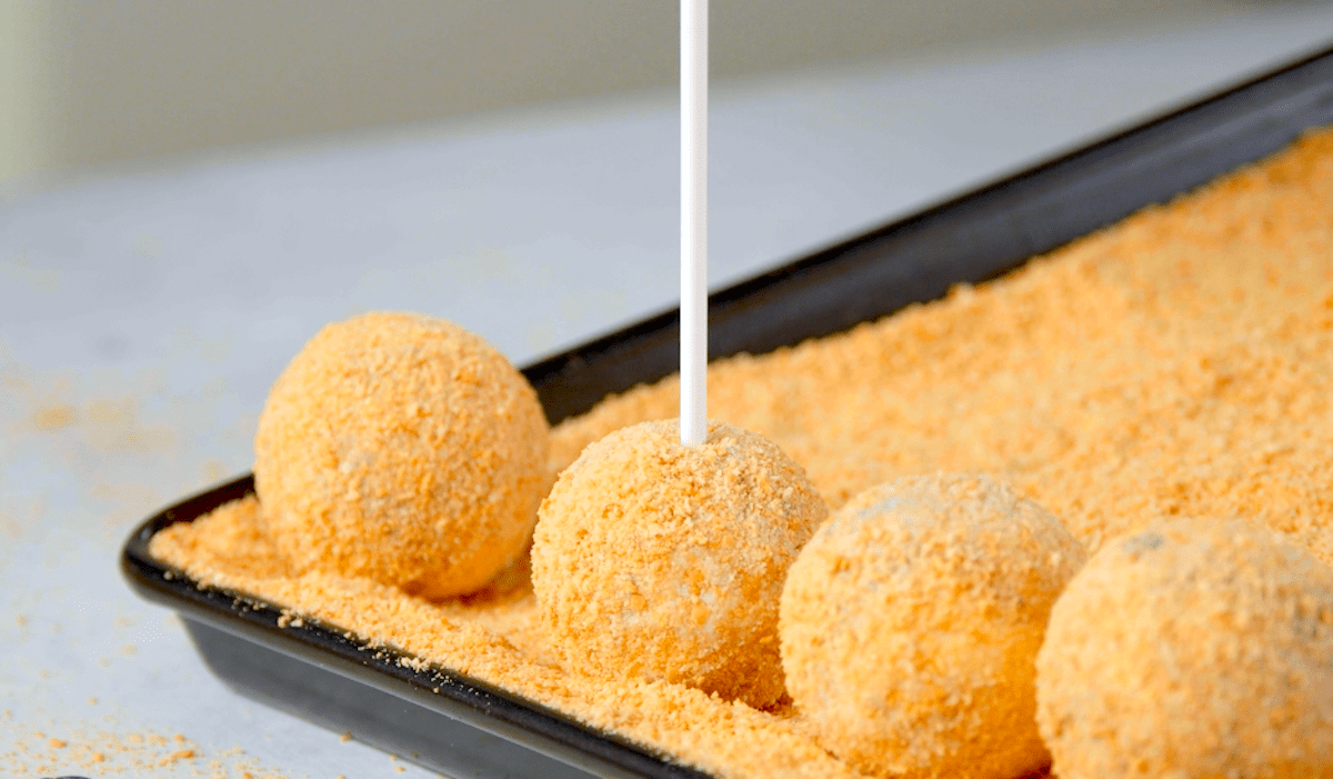 Ein Cake-Pop-Stiel wird in mit KekskrÃ¼meln ummantelte FrischkÃ¤sebÃ¤llchen gedrÃ¼ckt.