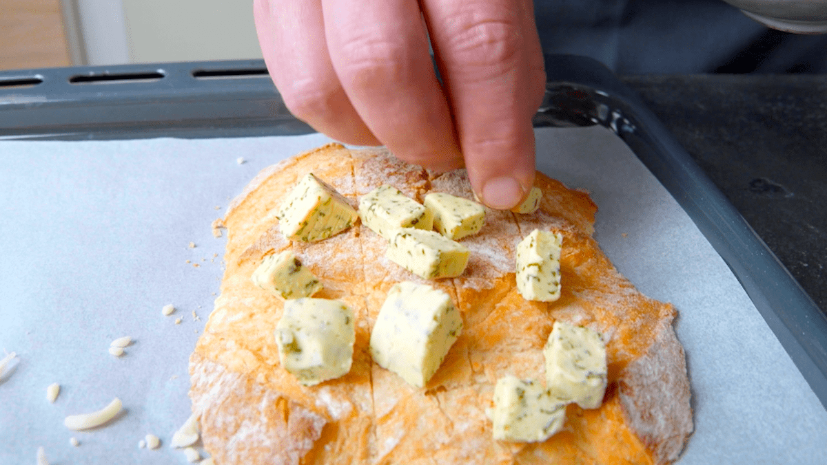 Mehrere StÃ¼ckchen KrÃ¤uterbutter werden auf einem StÃ¼ck WeiÃŸbrot verteilt.