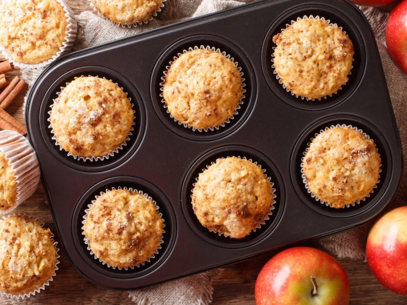 Ein Muffinblech mit Apfel-Zimt-Muffins. Daneben liegen Äpfel und weitere Muffins.