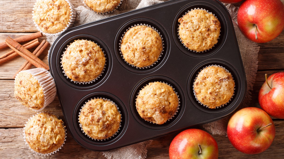 Ein Muffinblech mit Apfel-Zimt-Muffins. Daneben liegen Ã„pfel und weitere Muffins.