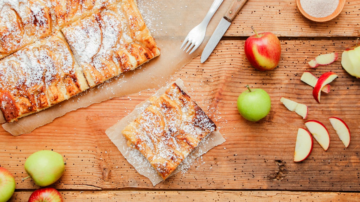 Ein StÃ¼ck Apfelkuchen vom Blech mit Puderzucker. Daneben Ã„pfel, Besteck und der restliche Kuchen.