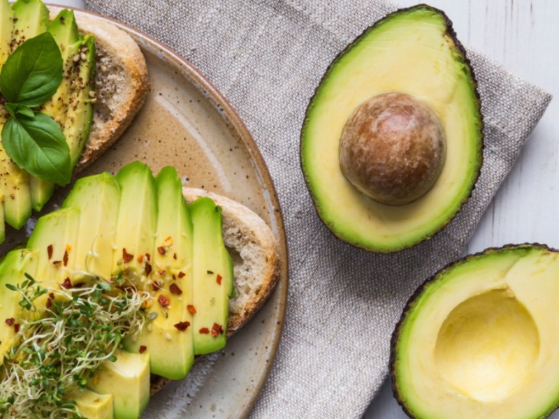 Eine halbierte Avocado neben zwei Broten mit Avocado auf einem Teller.