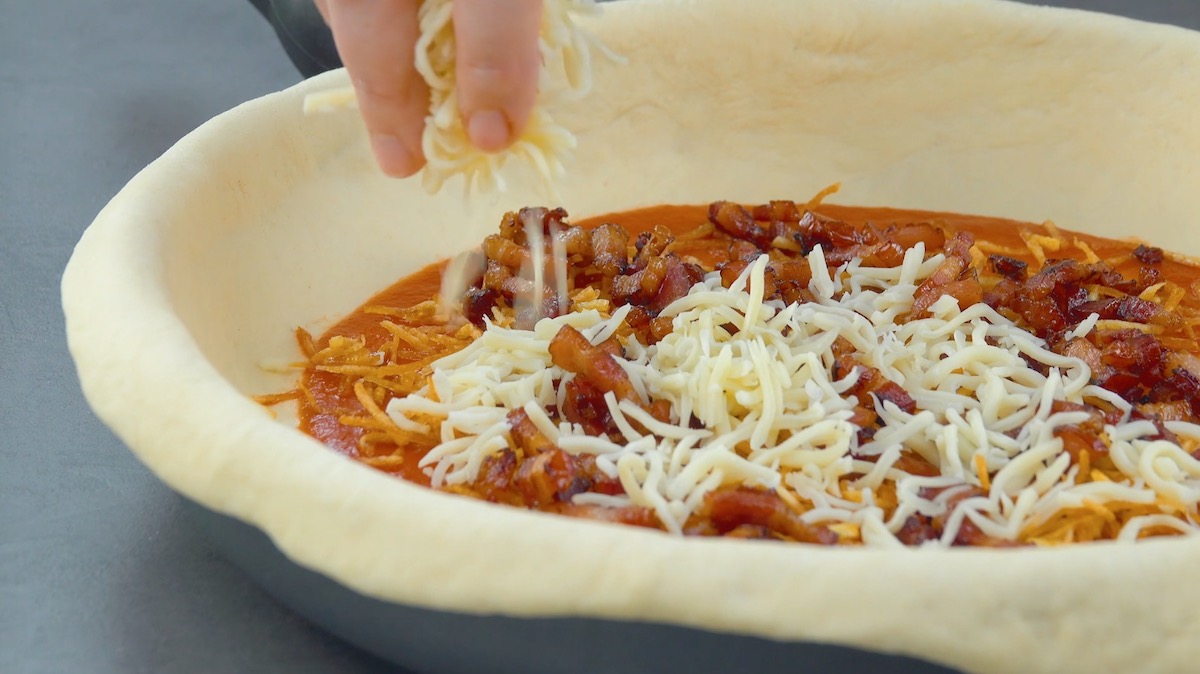 Pizzateig in Pfanne wird mit Tomatensauce, Kartoffelstiften, BaconwÃ¼rfeln sowie geriebenen Mozzarella belegt