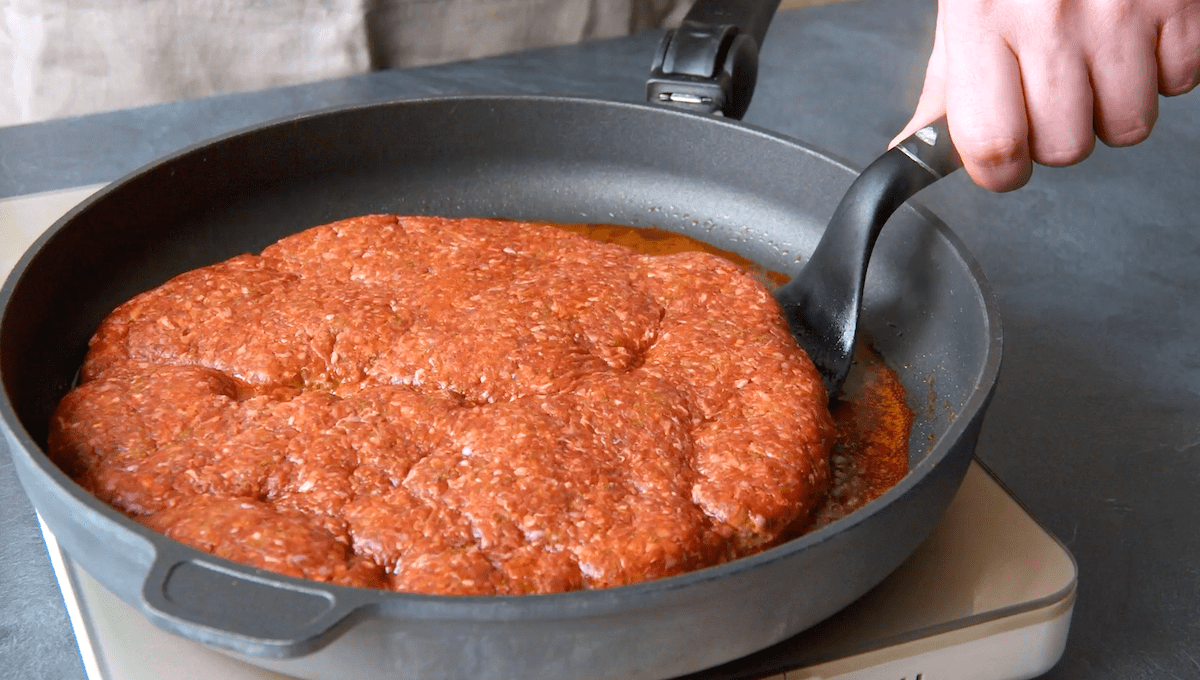 Ein pizzagroÃŸer Hackfleisch-Patty wird in einer Pfanne angebraten.