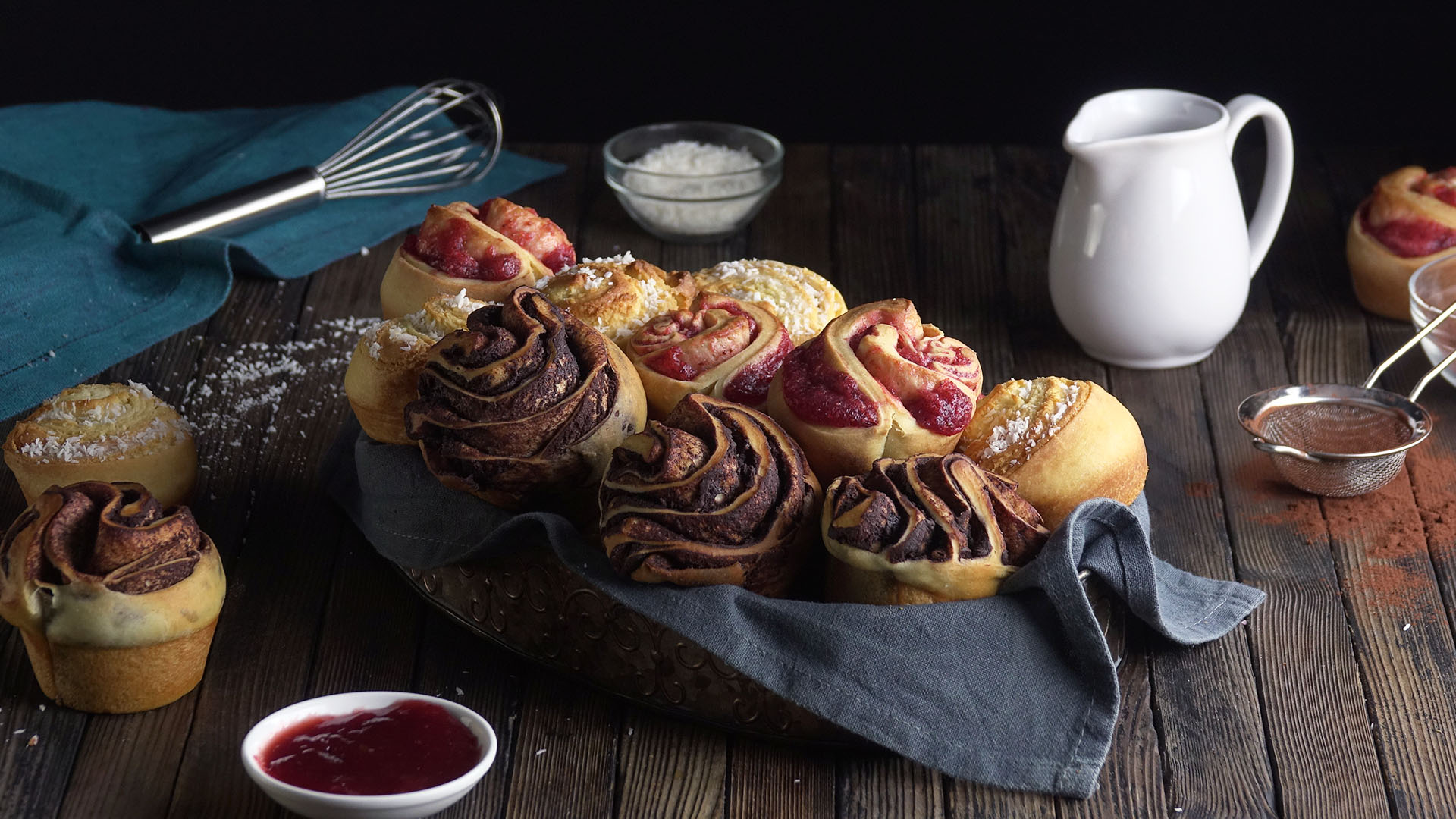 Drei Sorten Cruffins in einem Brotkorb mit Backutensilien, Schalen mit Backzutaten und einem Kännchen im Hintergrund. 