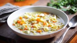 Ein Teller Hühnersuppe mit Nudeln. Daneben ein Löffel und ein Geschirrtuch. Im Hintergrund frische Petersilie.