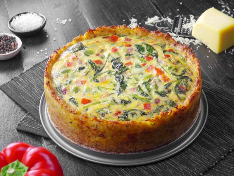 Kartoffel-Quiche auf einem Teller, darunter liegen zwei Tischsets. Drum herum liegen Paprika, Butter, Salz und Pfeffer als Dekoration.