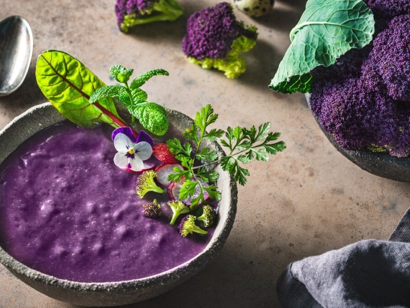 Lilafarbene Süßkartoffel-Blumenkohl-Suppe mit Salat-Dekoration. Daneben liegt ein Löffel und im Hintergrund Zutaten.