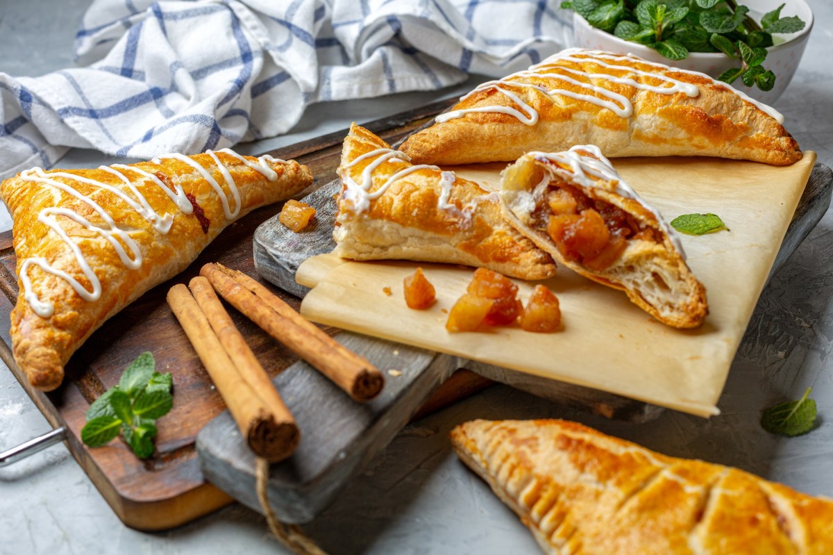 Ein paar Apfel-Kürbis-Taschen auf einem Brett, einige davon angebrochen, daneben Zimtstangen und ein Geschirrtuch.