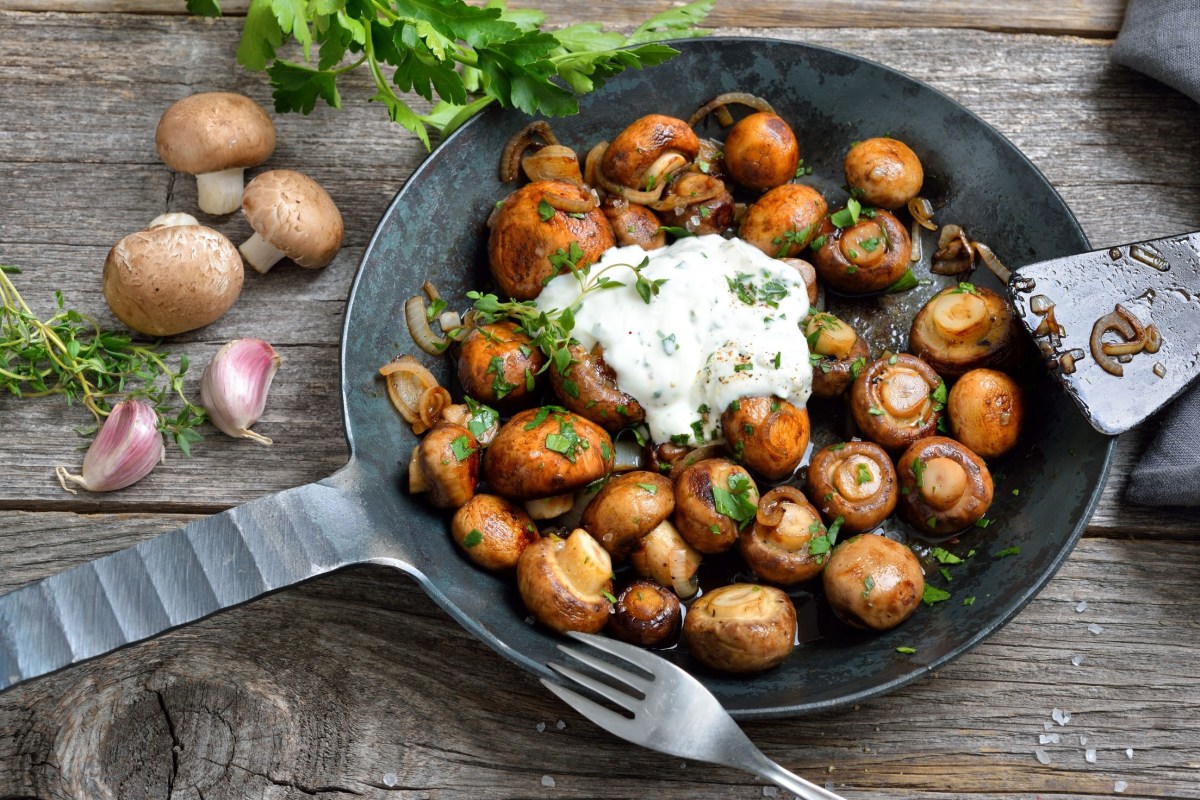 Gusseiserne Pfanne gefüllt mit Champignons wie vom Weihnachtsmarkt. Ein Klecks Knoblauchsoße in der Mitte. Pilze und Kräuter als Deko im Hintergrund.