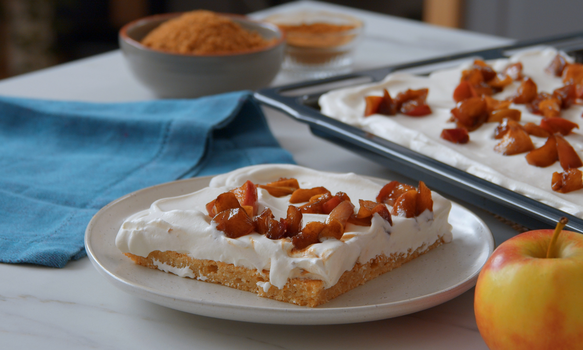 Ein Stück Apfelkuchen neben dem Blech mit Kuchen und einer Schüssel Zimt im Hintergrund.