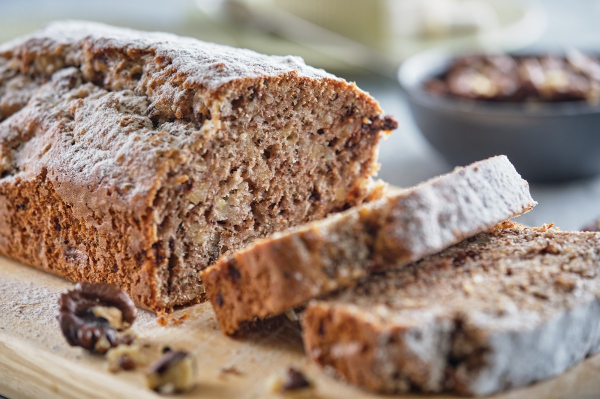 Ein angeschnittenes einfaches Dinkel-Vollkornbrot auf einem Brett, auf dem auch Walnüsse liegen, im Hintergrund unscharf Schüsseln mit Nüssen.