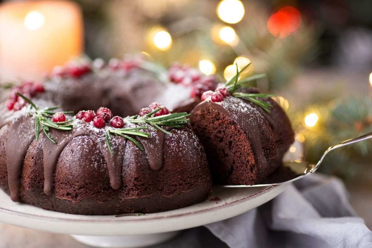 Ein angeschnittener Glühwein-Gugelhupf, von dem mit einem Tortenheber ein Stück herausgenommen wird, im Hintergrund unscharf Lichterketten.