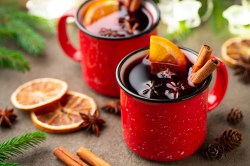 2 rote Tasse mit Glühwein, der mit Glühweingewürzen wie Zimt und Anis sowie Orangenscheiben garniert ist.