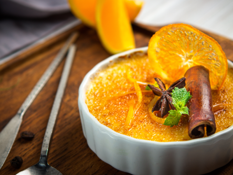 Ein überbackener Orangenpudding mit Minze, Zimtstange und Orangenscheibe in Schälchen auf Holzbrett, im Hintergrund eine angeschnittene Orange.