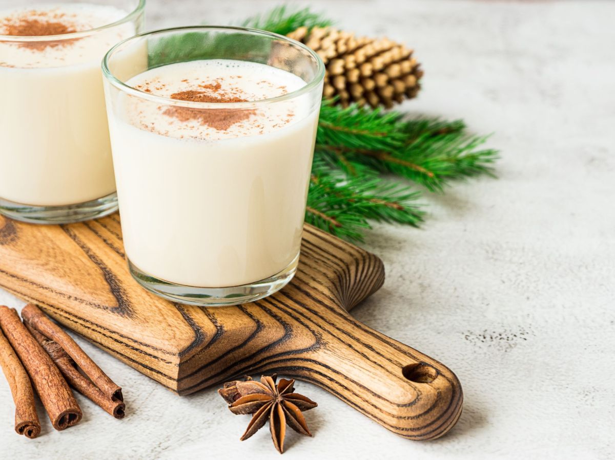 Vanille-Gewürz-Smoothie, serviert in zwei Gläsern, dekoriert mit Kakaopulver. Die Gläser stehen auf einem Holzbrett. Vanilleschote und Gewürze liegen als Deko daneben, im Hintergrund sind Tannenzweige zu sehen