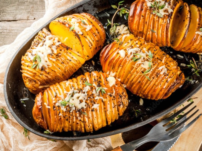 Vier Akkordeon-Kartoffeln in einer schwarzen Auflaufform mit Kräutern und Käse.