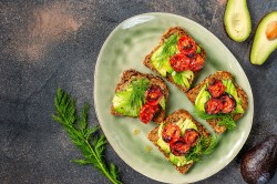 3 Avocado-Brote mit Ofentomaten und Kräutern belegt auf einem Teller in der Draufsicht, drumherum ganze und halbierte Avocados.