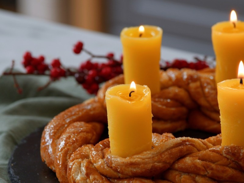 Ein essbarer Adventskranz aus Brezelteig mit vier Kerzen aus Butter. Im Hintergrund eine grüne Serviette und rote Pflanzen, passend zu Weihnachten.