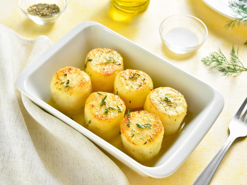 Eine weiße rechteckige Auflaufform mit Fondant-Kartoffeln, bestreut mit Rosmarin, daneben eine Gabel und ein paar Zutaten wie Brühe und flüssige Butter.