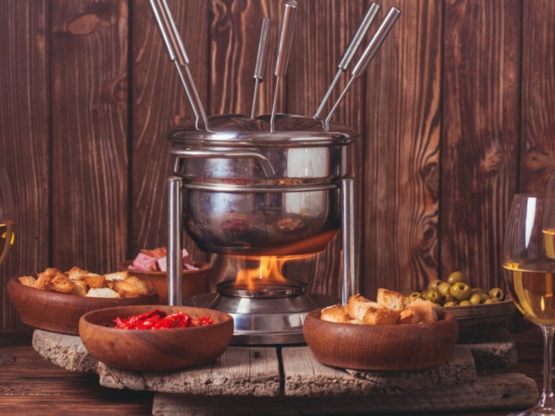 Ein Fondue mit unterschiedlichen Zutaten drumherum. Daneben ein Glas Weißwein.