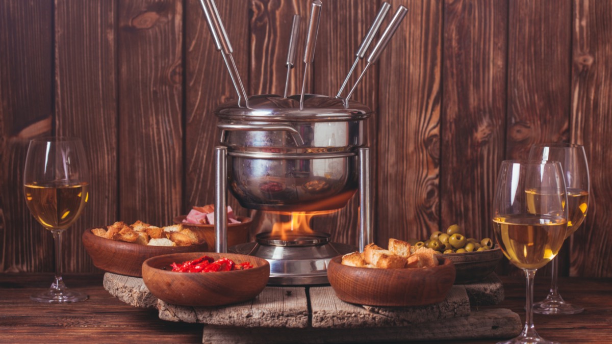 Ein Fondue mit unterschiedlichen Zutaten drumherum. Daneben ein Glas Weißwein.