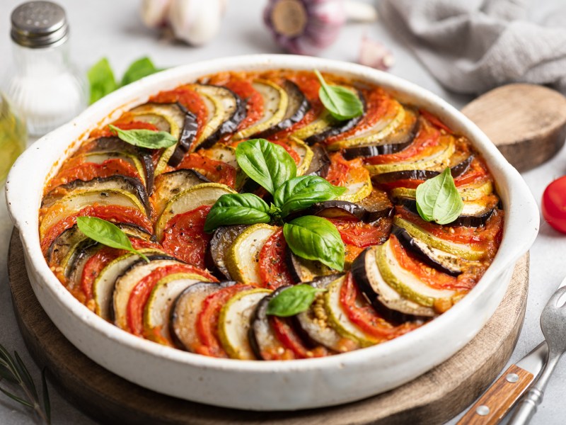 französischer Gemüseauflauf Tian de Légumes in einer runden Auflaufform auf einem Holzbrett, drumherum viele Zutaten.