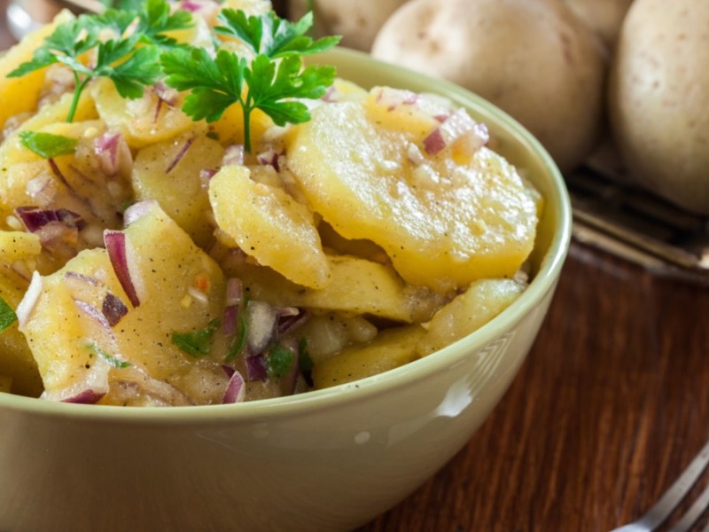 Eine Schüssel mit Kartoffelsalat, garniert mit Petersilie. Daneben liegt eine Gabel und rohe Kartoffeln im Hintergrund.