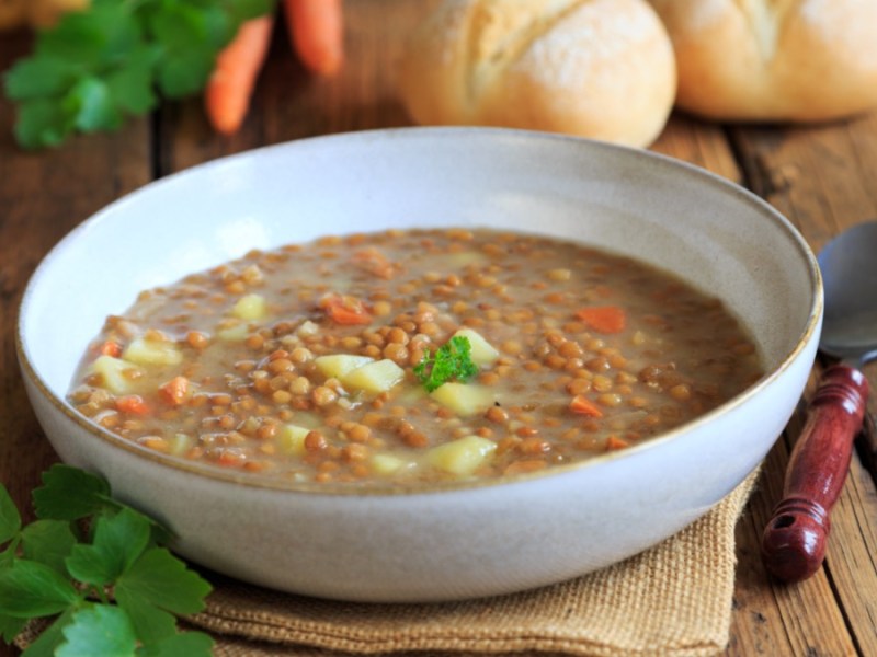 Eine Schüssel mit Omas Linsensuppe. Daneben liegen ein Löffel und einige der Zutaten.