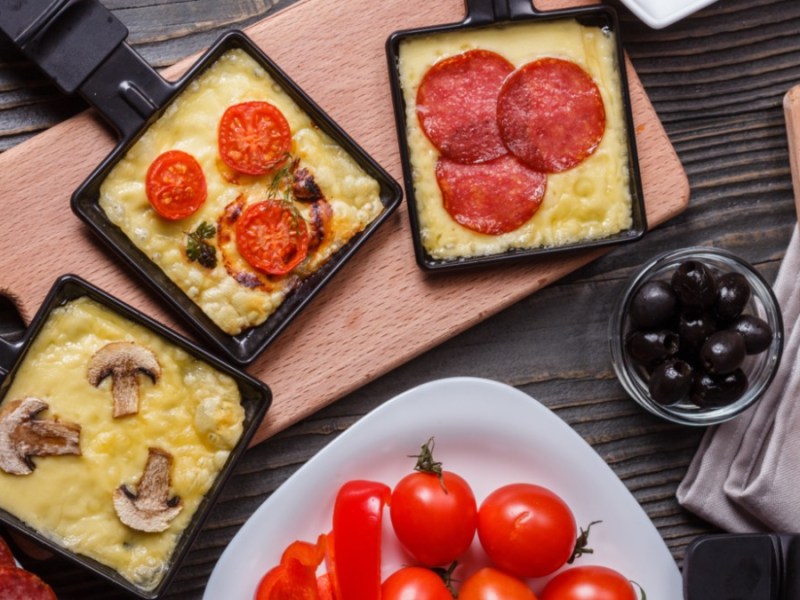 Drei Raclette-Pfännchen, die unterschiedlich gefüllt sind, in der Draufsicht. Drumherum liegen Tomaten und weitere Zutaten.