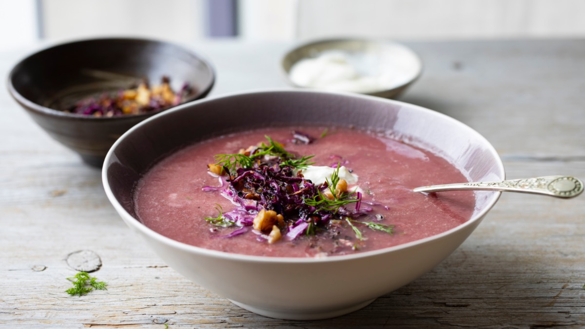 Ein Teller Rotkohlsuppe, in der ein Löffel liegt. Daneben zwei Schalen, eine mit Sojacreme und eine mit Topping aus Walnüssen und Rotkohl.