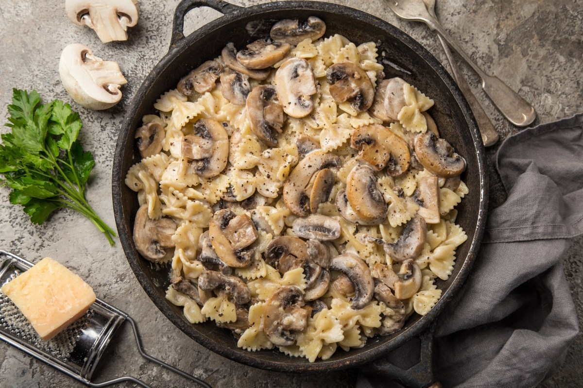 Eine gusseiserne Pfanne mit Walnuss-Pilz-Pasta in der Draufsicht, daneben ein Stück Parmesan und ein paar Champignons.