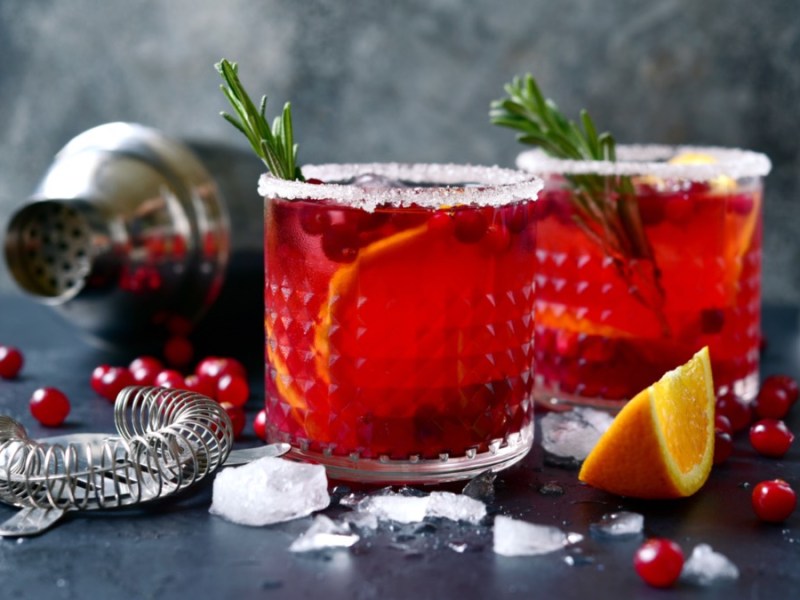 2 Gläser Winter-Margarita mit Rosmarinzwigen. Im Hintergrund ein Cocktailshaker, Eiswürfel, Cranberrys, Orangen und Rosmarinzweige.