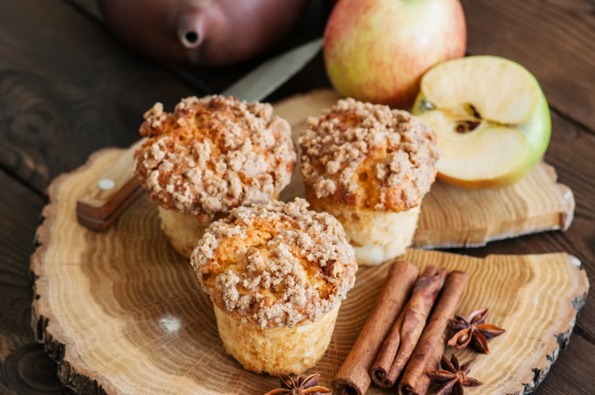 3 Apfel-Streusel-Muffins auf einer runden Holzplatte, daneben Äpfel und Zimtstangen.