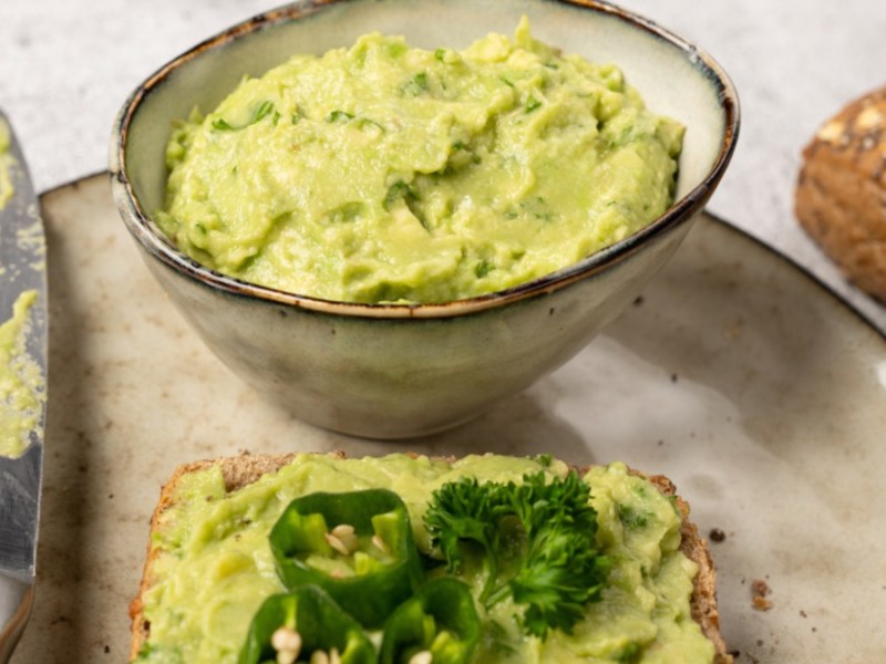 Ein großer Teller mit einer Schale Avocado-Cashew-Aufstrich, einer Scheibe Brot mit Aufstrich und einem Messer. Im Hintergrund liegt ein Vollkornbrötchen.