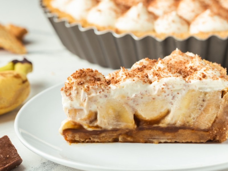 Ein Stück Banoffee Pie auf einem Teller. Daneben liegen Banane und der Rest der Pie ist im Hintergrund zu sehen.