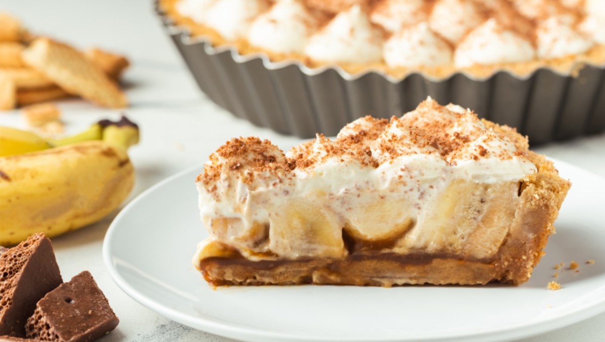 Ein Stück Banoffee Pie auf einem Teller. Daneben liegen Banane und der Rest der Pie ist im Hintergrund zu sehen.