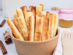 Blätterteig-Pudding-Stangen, angerichtet in einer Schale. Daneben liegen Zimtstangen und ein Holzschaber als Dekoration. Im Hintergrund ist ein Marmeladenglas zu sehen.