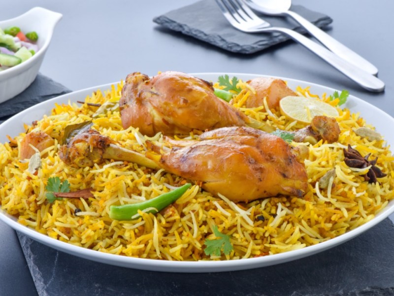 Ein tiefer Teller mit Chicken Biryani. Im hintergrund sind Besteck und eine Schale mit Salat.