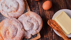 Drei Ensaimadas auf einem Holzbrett. Daneben liegen ein Ei und ein Teller mit einem Stück Butter und einem Holzlöffel darauf.