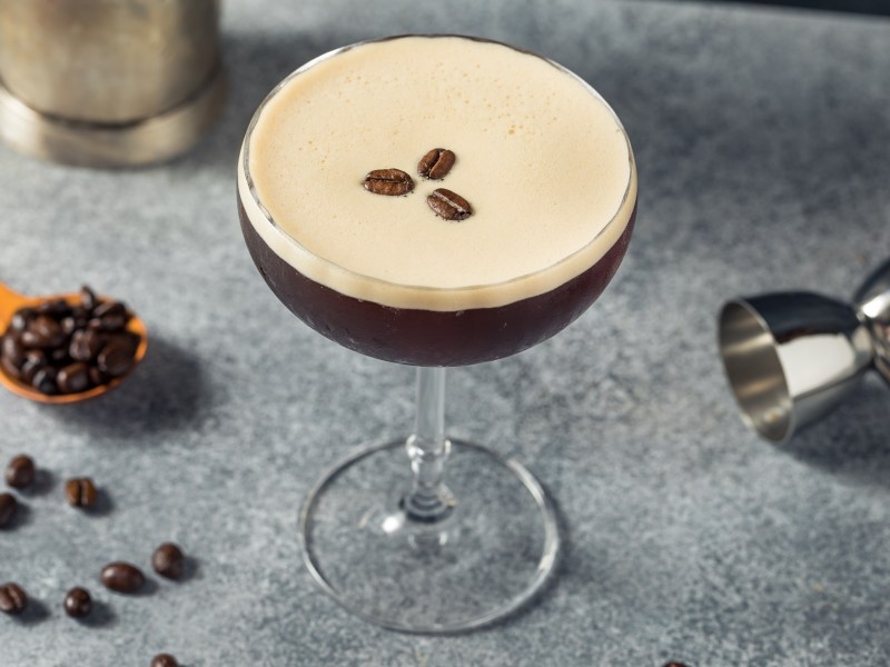 Ein Glas Espresso Martini mit drei Kaffeebohnen garniert, daneben Kaffeebohnen und Cocktailzubehör