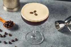 Ein Glas Espresso Martini mit drei Kaffeebohnen garniert, daneben Kaffeebohnen und Cocktailzubehör