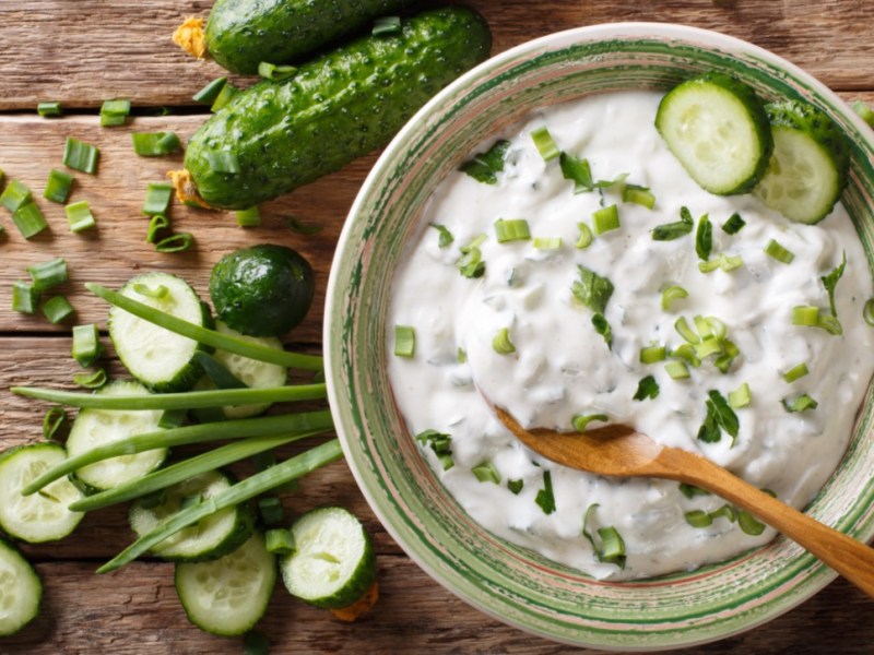 Eine Schüssel mit Gurken-Raita, in dem ein Holzlöffel liegt. Daneben liegen Gurken und frische Zutaten.