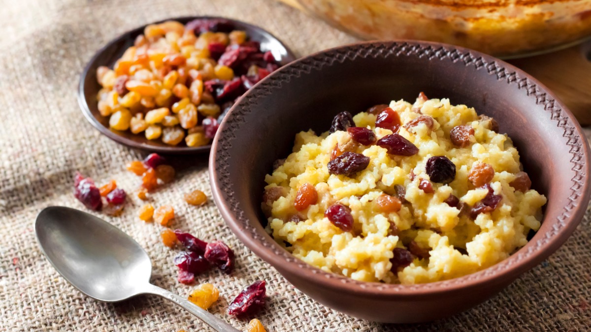 Eine Schüssel mit süßen Hirsebrei mit Rosinen und Cranberrys. Daneben liegt ein Löffel und eine kleine Schale mit Rosinen.